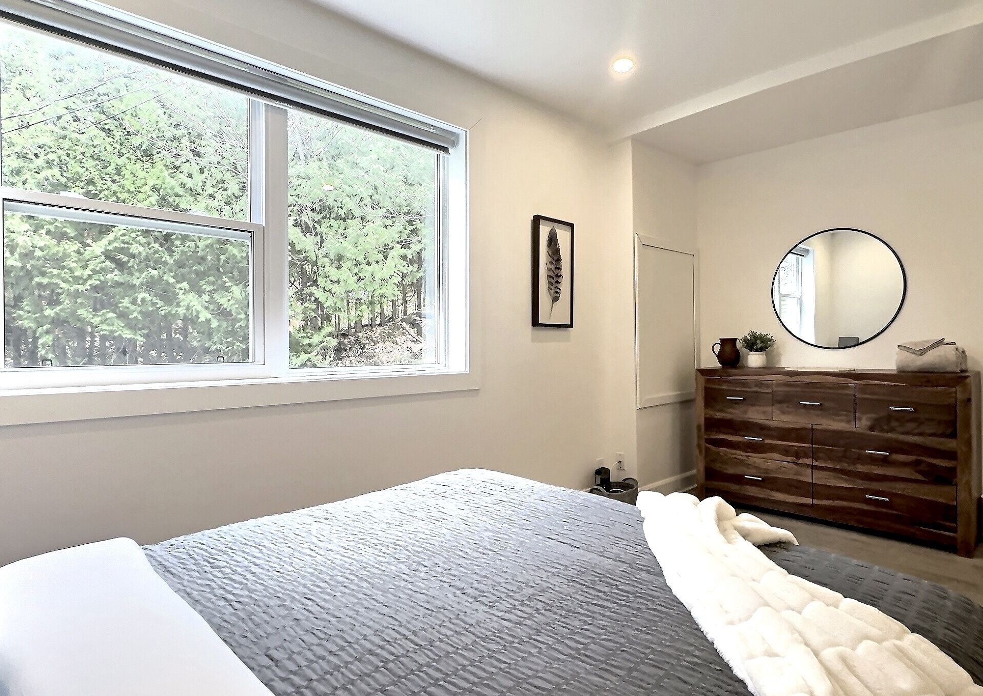 A bedroom with a large window, a bed with a textured blanket, a dresser with drawers, a mirror, and a framed picture on the wall.