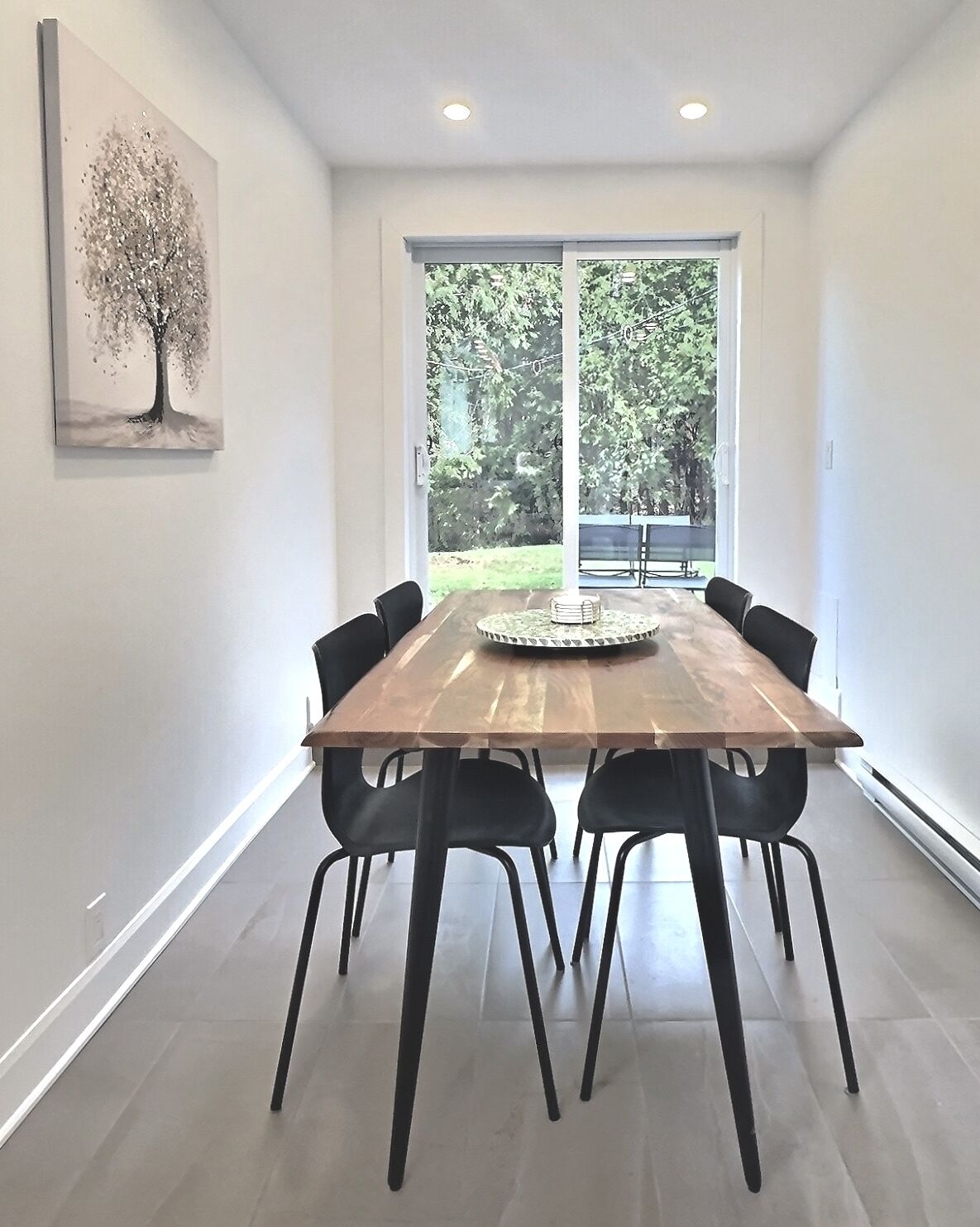 Een moderne eetruimte met een houten tafel en zwarte stoelen, grote ramen met uitzicht op groen, en een schilderij aan de muur.
