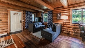 Interior de cabana de madeira com teto de madeira, um sofá, um pufe, uma mesa pequena e uma janela com vista para vegetação.