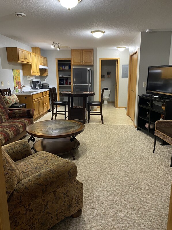 A living room with a sofa, a coffee table, and a TV. There is a kitchen area with cabinets and a refrigerator.