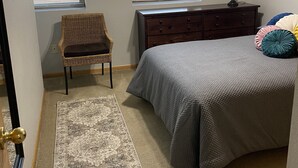 Ein Schlafzimmer mit Bett, Kommode, Stuhl und zwei Fenstern mit Jalousien.