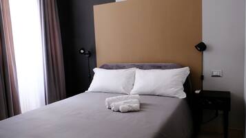 Una cama bien hecha con sábanas blancas y una toalla doblada, una mesita de noche negra con una lámpara, y vista a una ventana con cortinas.