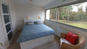 Une chambre avec un grand lit, une fenêtre donnant sur des arbres, un fauteuil en osier avec un coussin rouge et un luminaire au plafond.