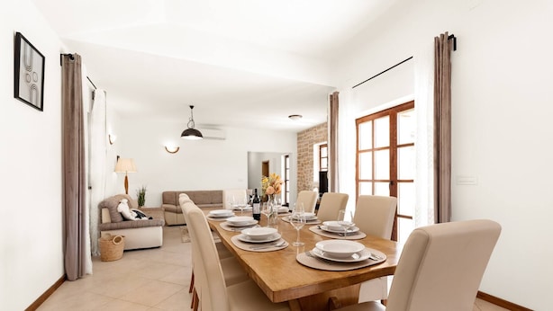 Un comedor con una mesa de madera dispuesta para cuatro personas, rodeada de sillas, y un sofá y una lámpara al fondo.