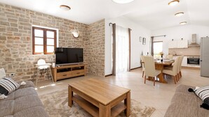 Un salón amplio con una pared de piedra, una mesa de centro de madera, un televisor sobre un mueble de madera, un comedor con mesa y sillas, y una cocina con armarios y un refrigerador.