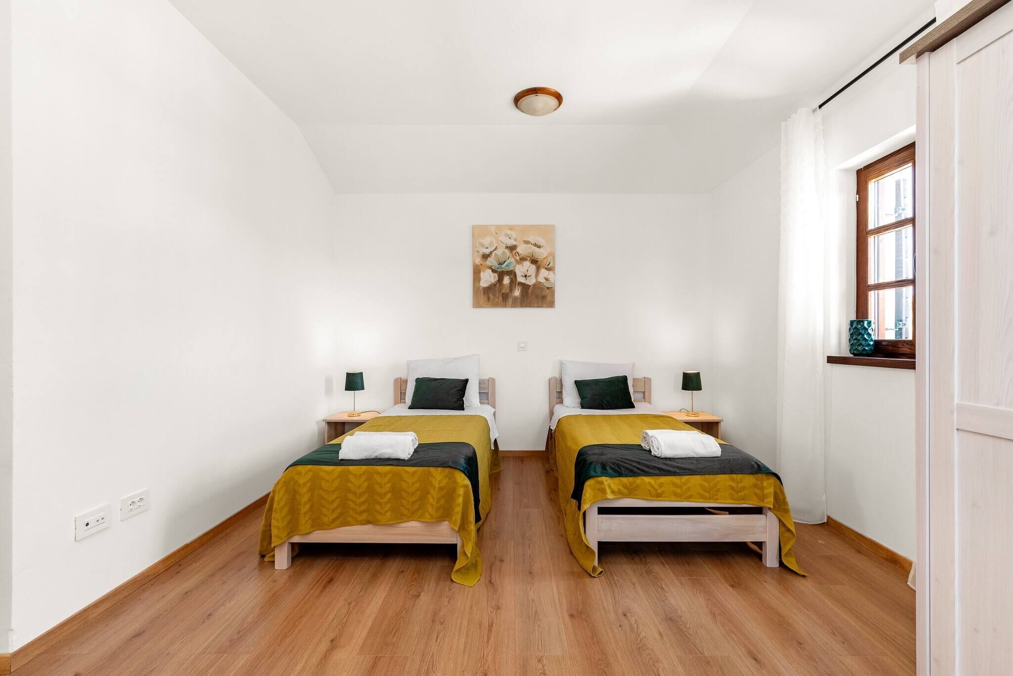 A room with two beds, wooden flooring, white walls, a ceiling light, a window with curtains, and a painting on the wall.
