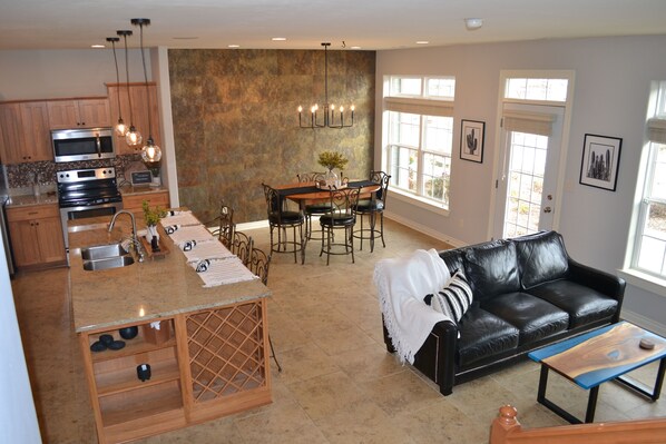 A modern kitchen with a central island, dining area with a table and chairs, a black leather sofa, and a window with a view.