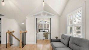 Ein modernes Wohnzimmer mit einer Holztreppe, einem grauen Sofa und Blick in ein Schlafzimmer durch eine Tür.