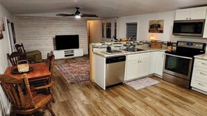 A compact kitchen with white cabinets, a built-in oven, microwave, and dishwasher. There's a dining area with a wooden table and chairs, and a TV mounted on the wall. - Country Living located Mid-Way between Granbury and Glen Rose.  (Granbury)