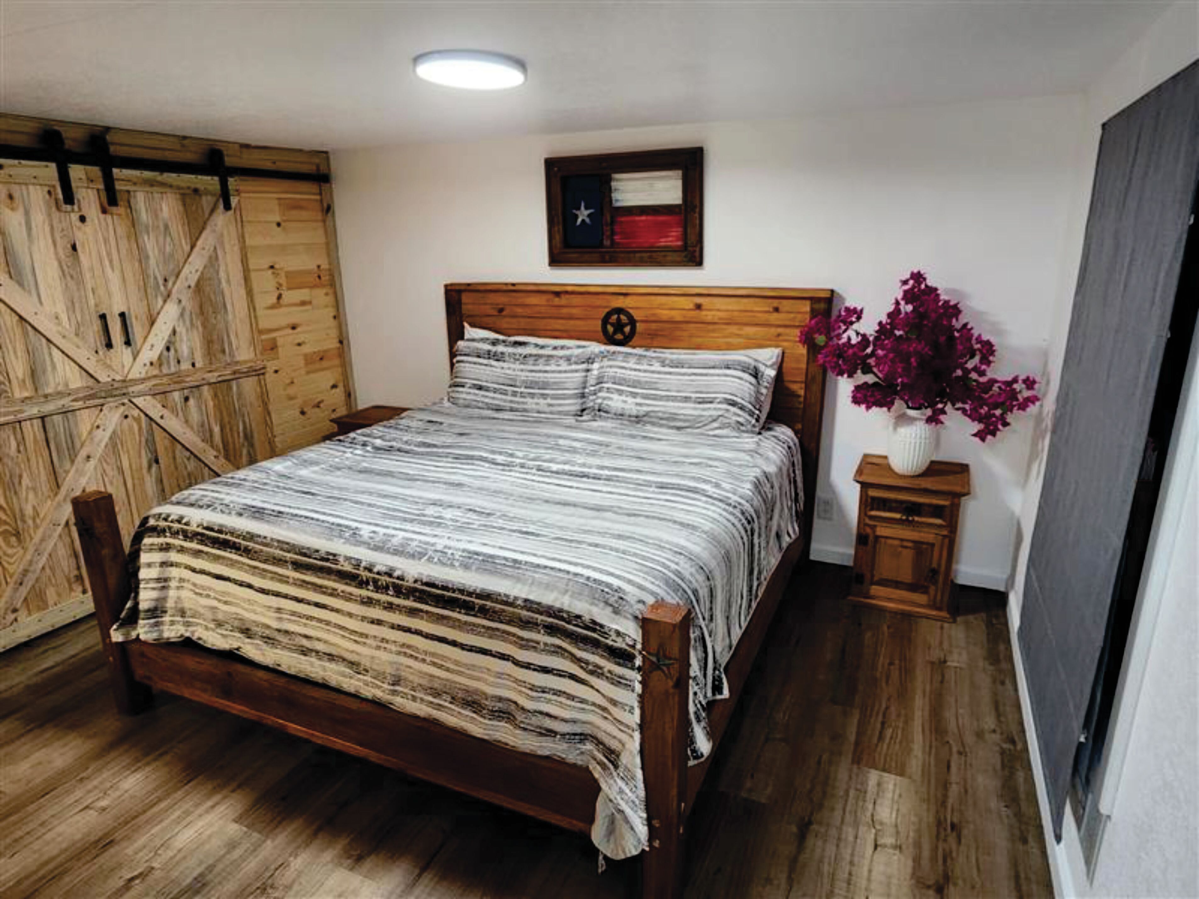 Een houten bed met een gestreept beddegoed, een houten hoofdbord met sterosdecoratie, een klein nachtkastje met een bloevas, en een houten wandpaneel.