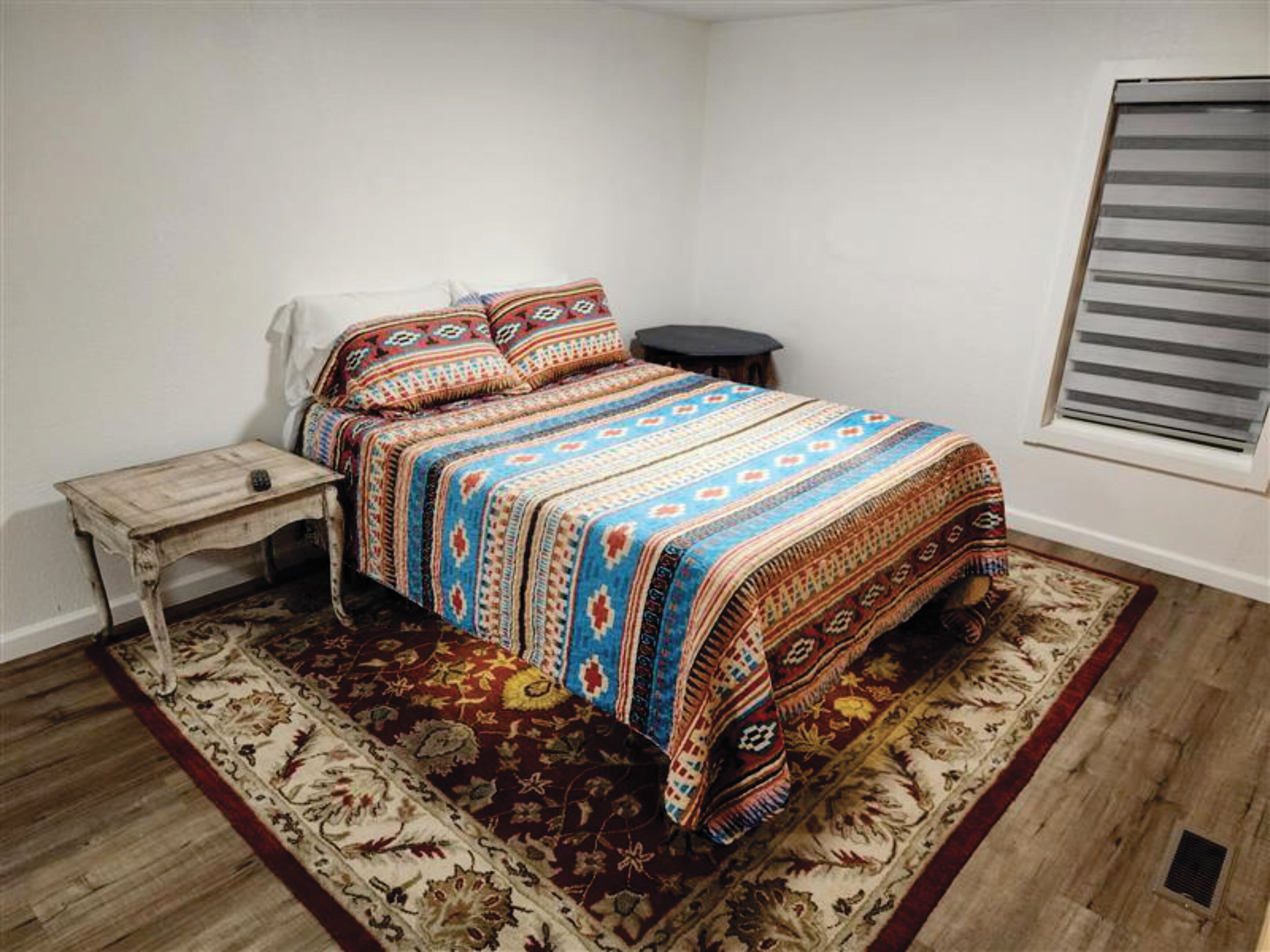 A bedroom with a bed, a nightstand, a small table, a window with blinds, and a patterned rug.