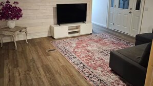 A living room with a flat-screen TV on a white stand, a patterned rug, a dark sofa, and a wooden floor. - Country Living located Mid-Way between Granbury and Glen Rose.  (Granbury)