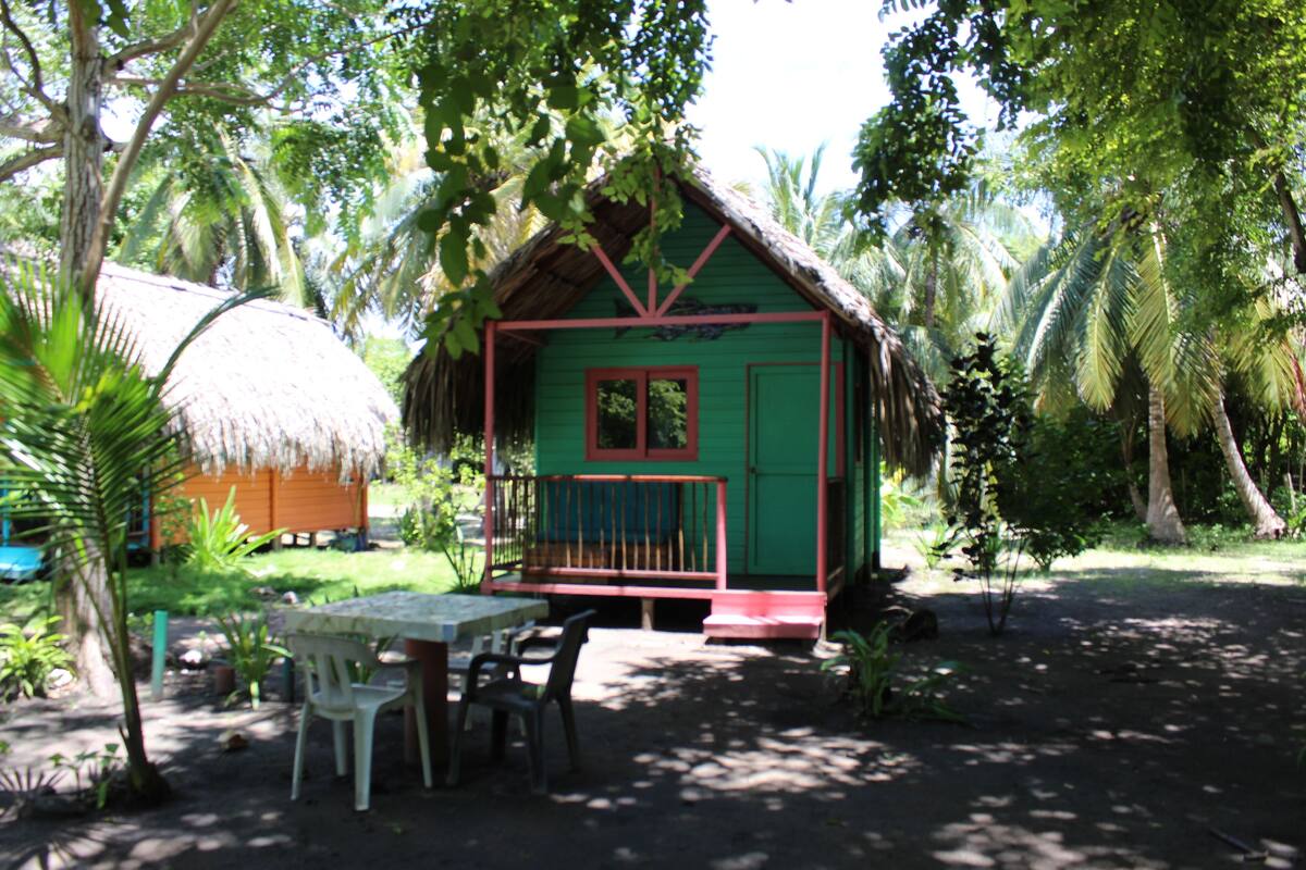 A small, green, thatched-roof hut with a red door and window, surrounded by lush greenery and palm trees.