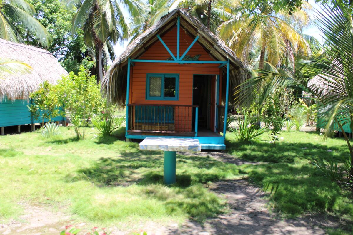A small, single-story house with a thatched roof, surrounded by lush greenery and palm trees.