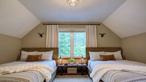A cozy bedroom with two beds, a window with curtains, a small table with a lamp, and wall-mounted deer antlers. - Baker Bliss | Hot Tub · Fire Pit · Pet-Friendly · Mt. Baker Gateway (Deming)