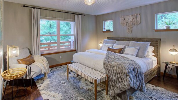 A bedroom with a large bed, a wooden headboard, a window with curtains, a wall-mounted macrame, and a floor lamp. - Baker Bliss | Hot Tub · Fire Pit · Pet-Friendly · Mt. Baker Gateway (Deming)