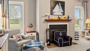Een woonkamer met een open haard, gestreept fauteuils, een blauwe fauteuil met een anker dessin, een schilderij van een zeilboot en een plank met decoratieve objecten.