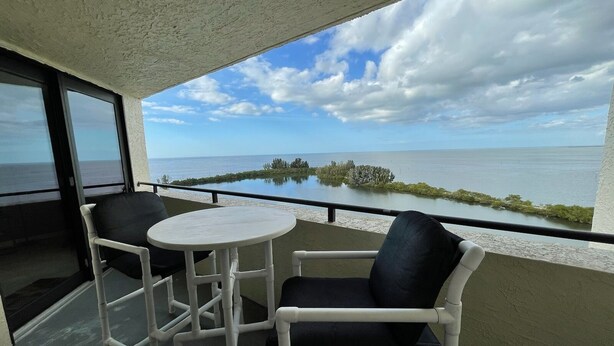 Un balcón con una mesa blanca y dos sillas, con vista al mar tranquilo y una pequeña isla.