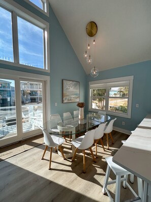 A modern dining area with a glass table and white chairs, large windows, and a view of the outside. - Ocean Haze - Oceanview Retreat (Carolina Beach)