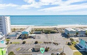 An aerial view of a coastal area with a parking lot, buildings, and a beach. - Ocean Haze - Oceanview Retreat (Carolina Beach)