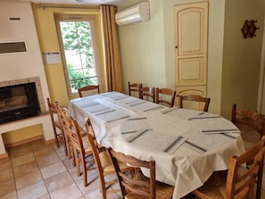Uma sala de jantar com uma mesa comprida coberta por um tecido branco, rodeada por cadeiras de madeira.