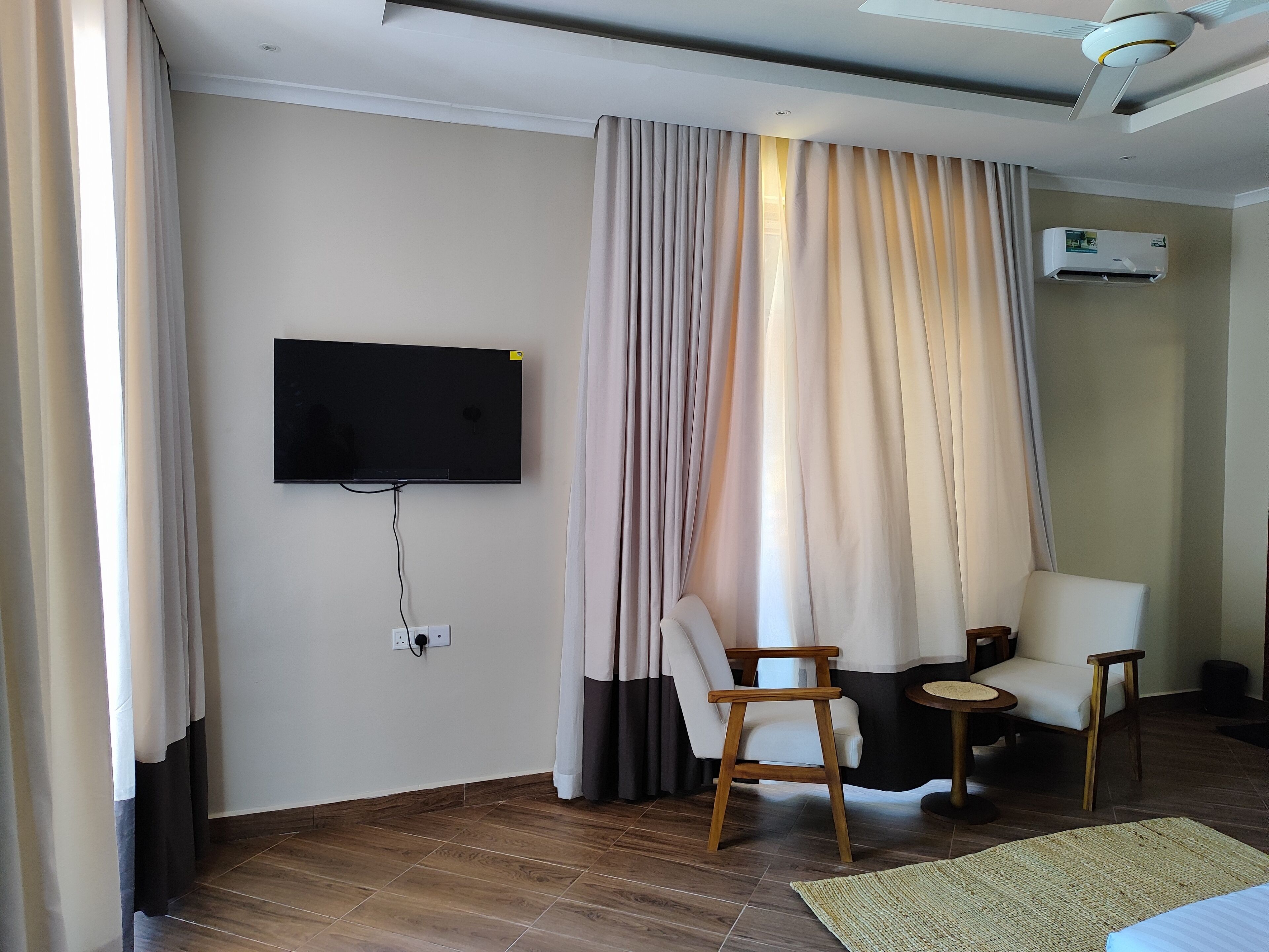 Ein Hotelzimmer mit Flachbildfernseher an der Wand, Holzboden, einem Bett, zwei Stühlen, einem kleinen runden Tisch und einem Deckenventilator.