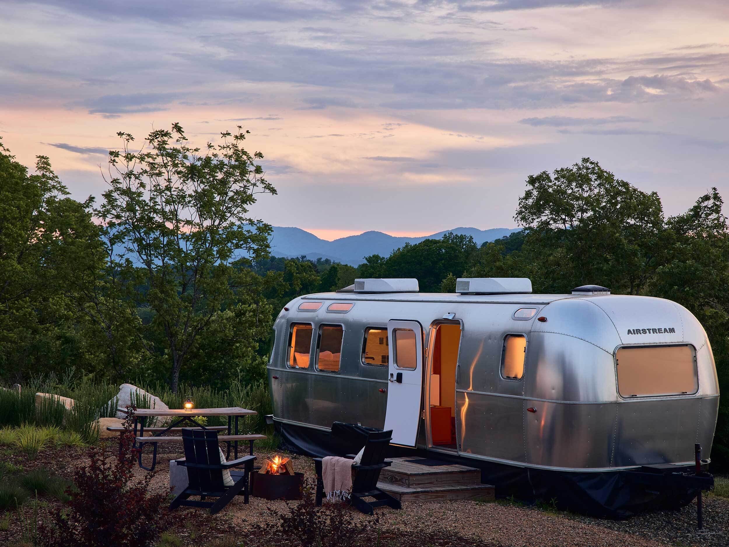 A silver AIRSTREAM trailer with a table and chairs, a fire pit, and a view of mountains.