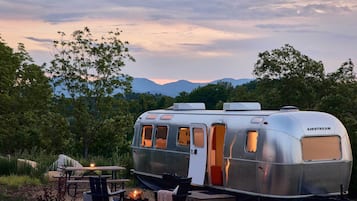 A silver AIRSTREAM trailer with a table and chairs, a fire pit, and a view of mountains.