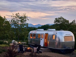 Een zilverkleurige AIRSTREAM caravan met een tafel, stoelen, een vuurkorf en uitzicht op bergen.