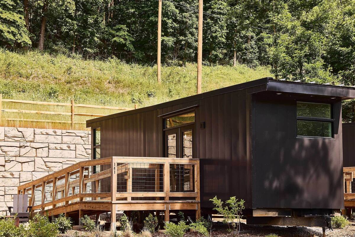 Una cabaña moderna con terraza de madera, pared de piedra y vista a una ladera boscosa.