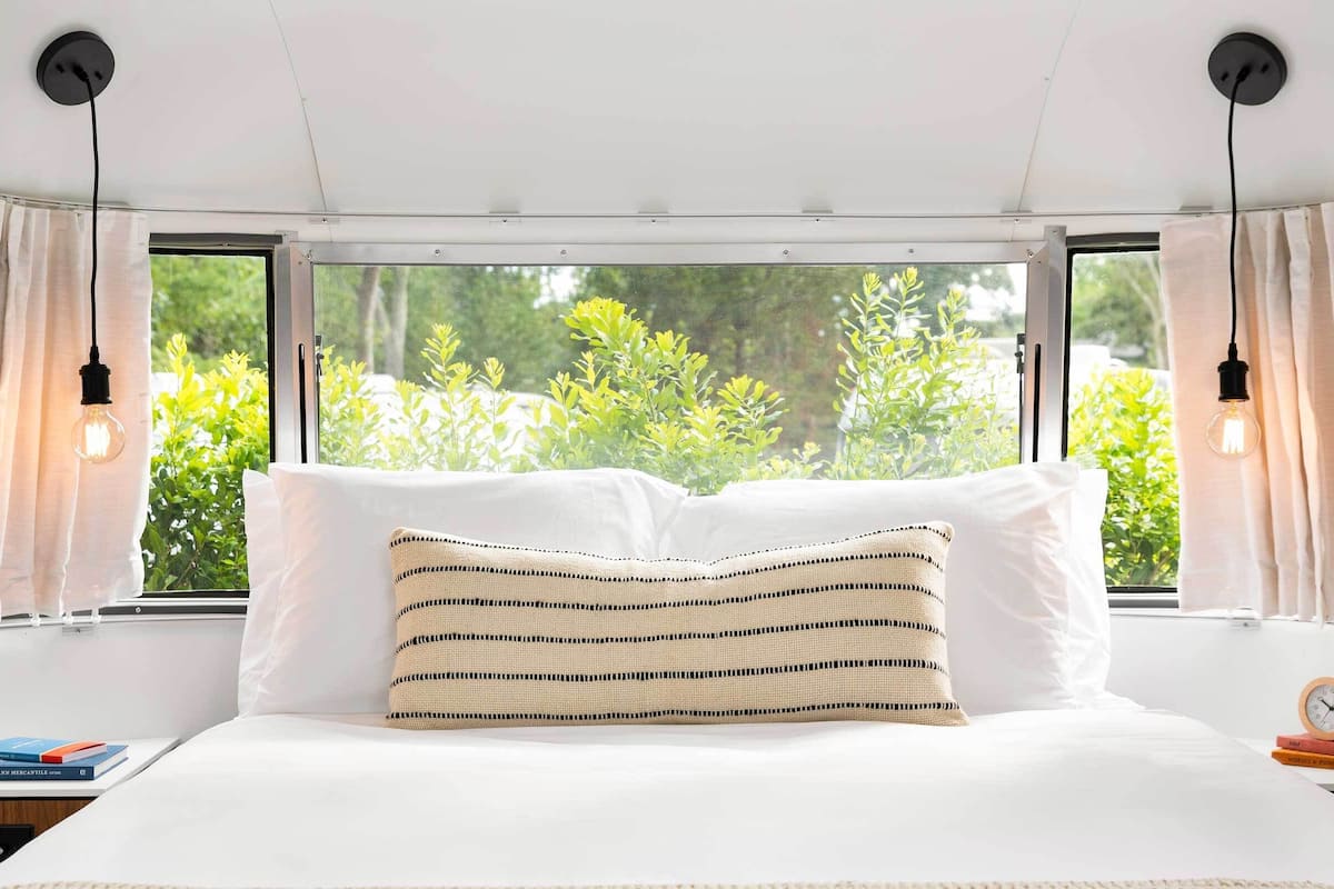 A bedroom with a large bed, two bedside tables, a window with curtains, and a view of greenery outside.