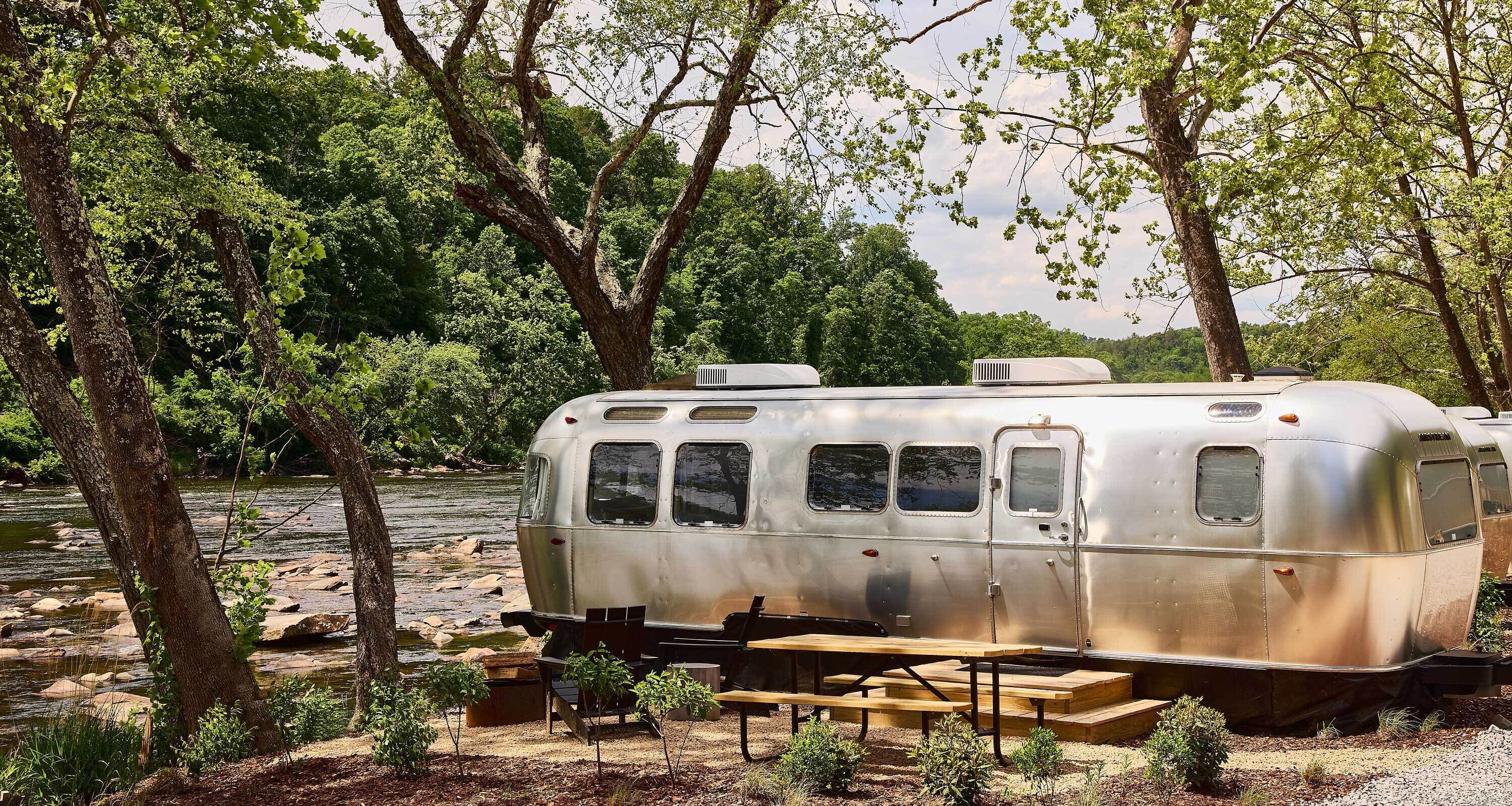 A vintage camper parked by a river with picnic tables and a small deck.