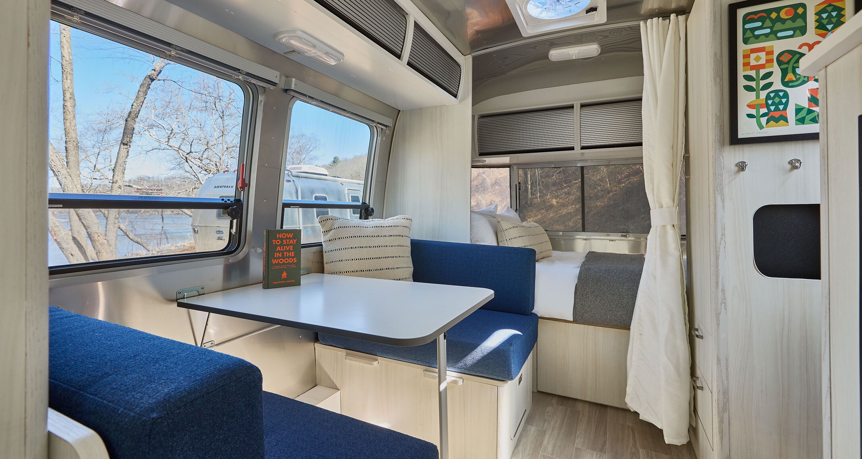 Interior compacto de autocaravana con una mesa, asientos azules, una pequeña cocina y una ventana que ofrece vistas a árboles y una caravana en el exterior.
