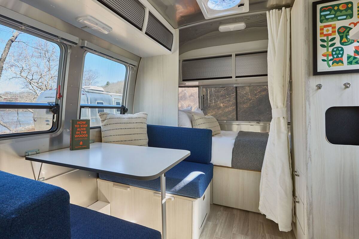 Interior compacto de autocaravana con una mesa, asientos azules, una pequeña cocina y una ventana que ofrece vistas a árboles y una caravana en el exterior.