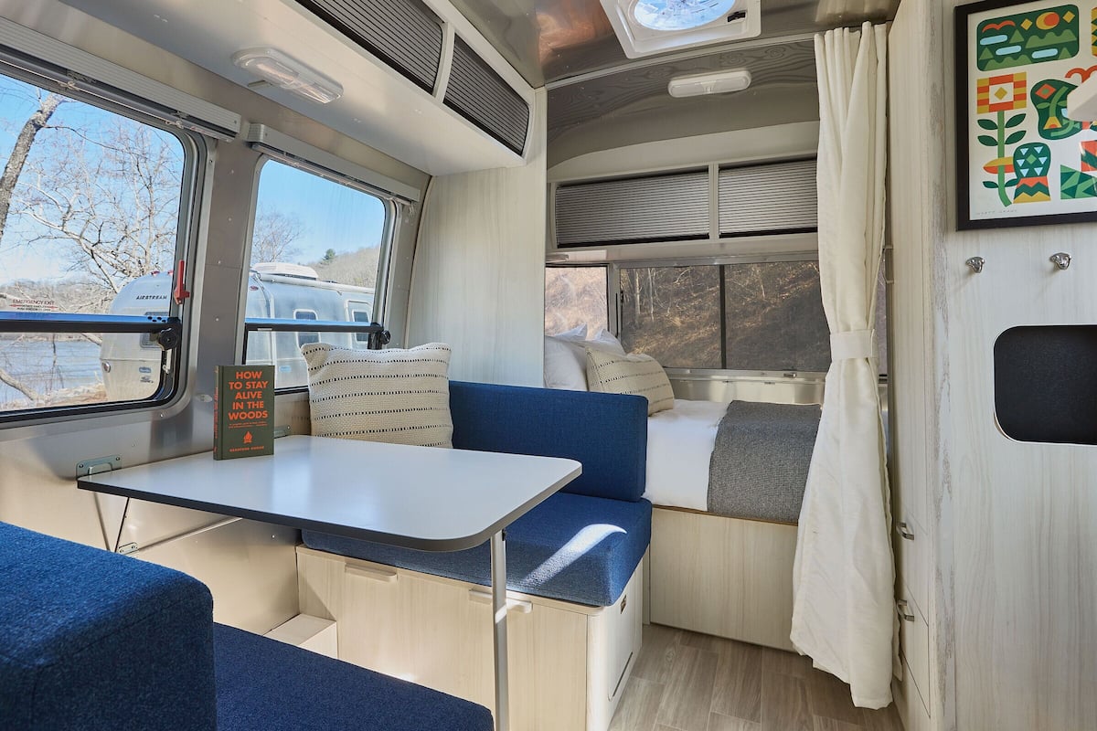 A compact RV interior with a table, blue seating, a small kitchenette, and a window with a view of trees and a trailer outside.
