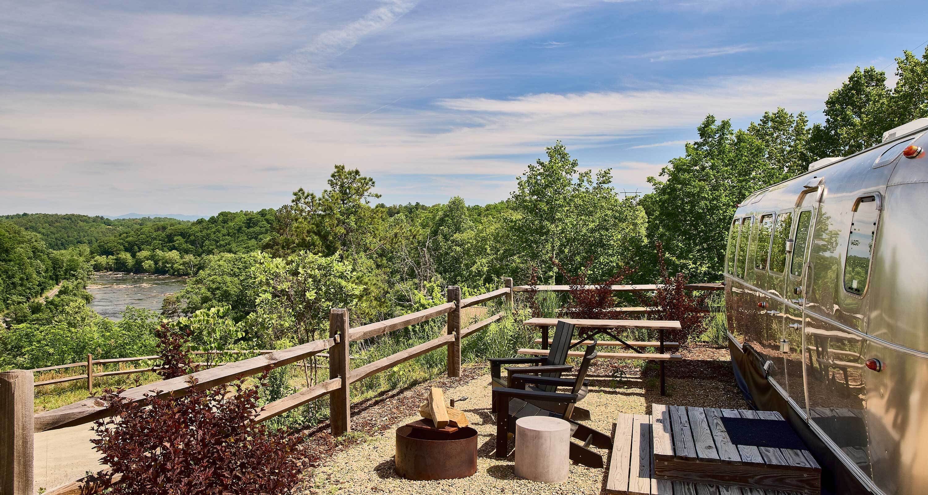 A rustic outdoor seating area with picnic tables and chairs, a wooden fence, and a view of a river and forest.