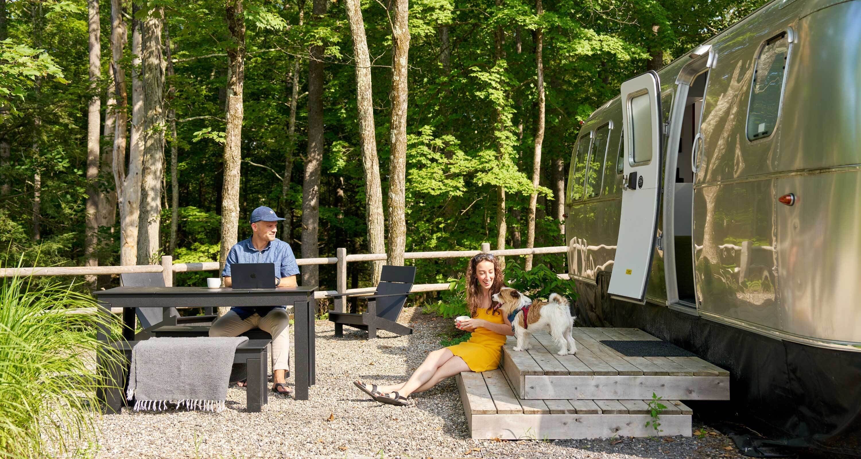 A forested area with a wooden deck, a table with chairs, and a trailer.