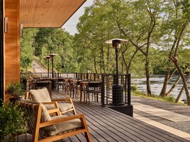 Eine Holzterrasse mit Sitzgelegenheiten im Freien, Blick auf einen Fluss und bewaldetes Gebiet.