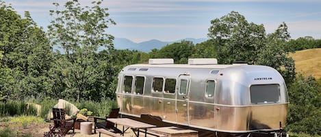 Un remolque plateado con kitchenette, un comedor con bancos y una mesa, y una vista panorámica de árboles y colinas al fondo.