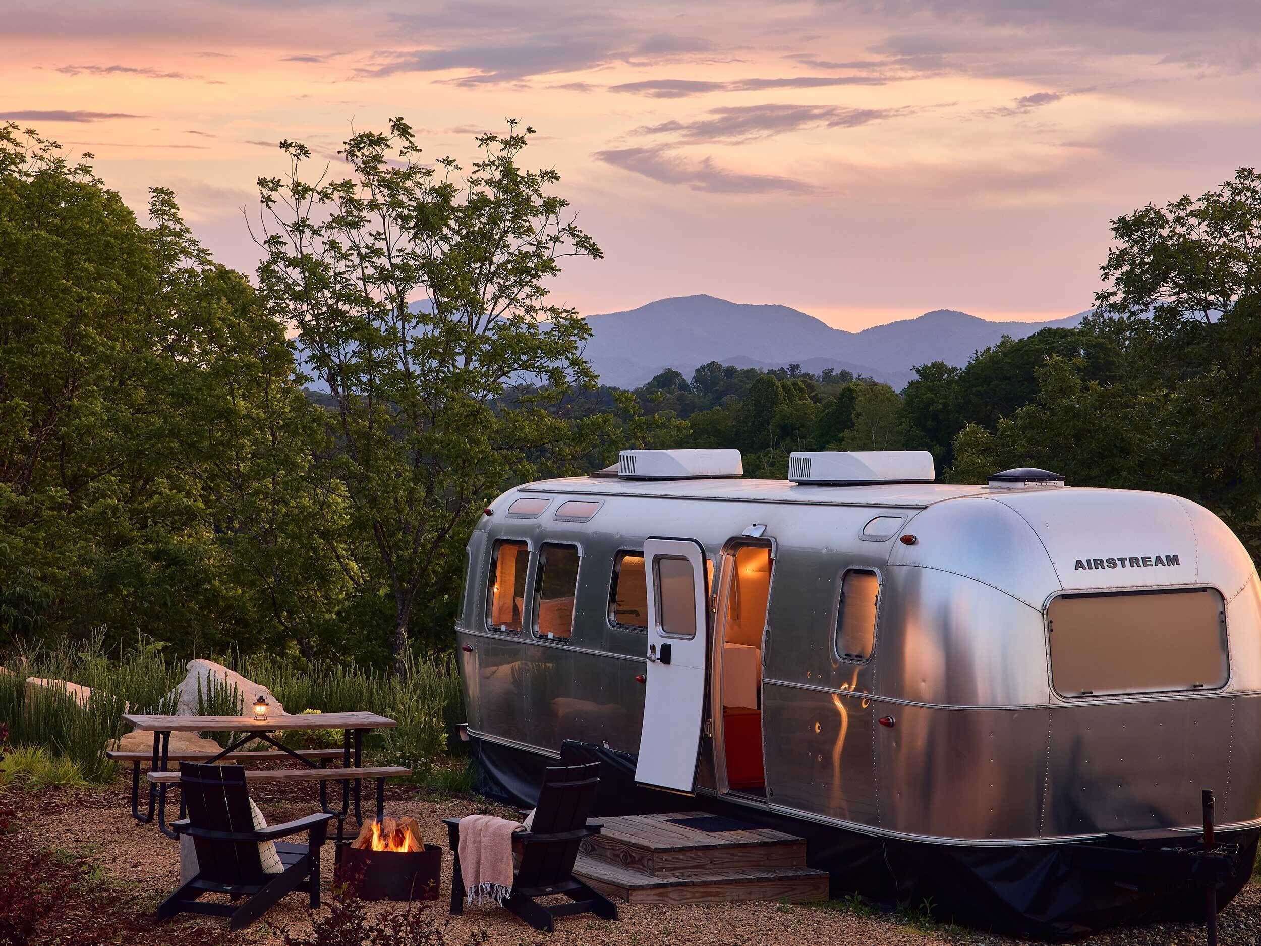 A silver trailer with a door open, a fire pit, and a picnic table with chairs.
