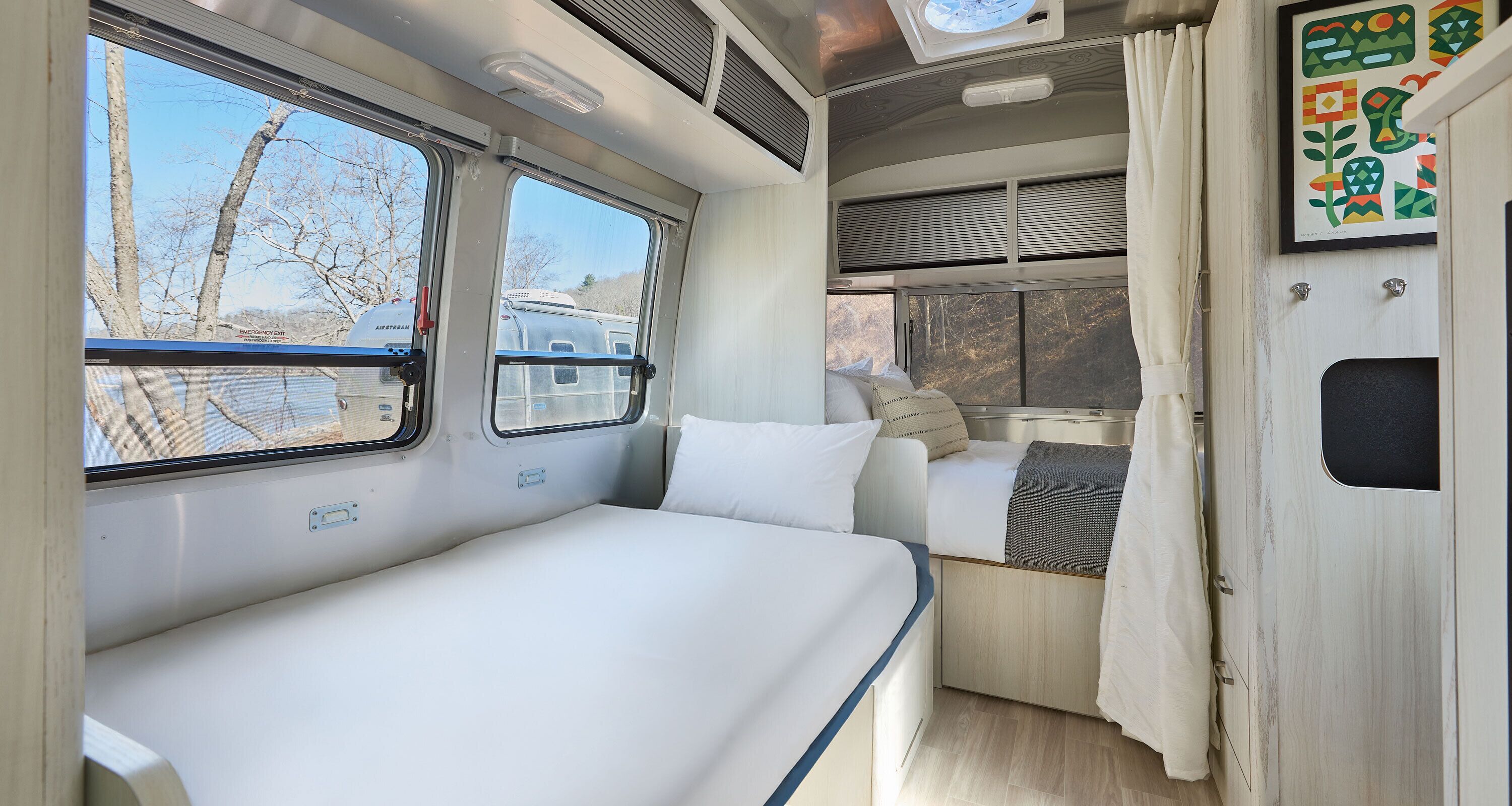 Interni di un camper compatto con un letto, una piccola zona salotto e una finestra che si affaccia su alberi e un rimorchio.