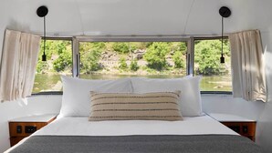 Ein Bett mit weißen und gestreiften Kissen, Blick auf einen Fluss und Grünflächen sowie ein Fenster mit Vorhängen.