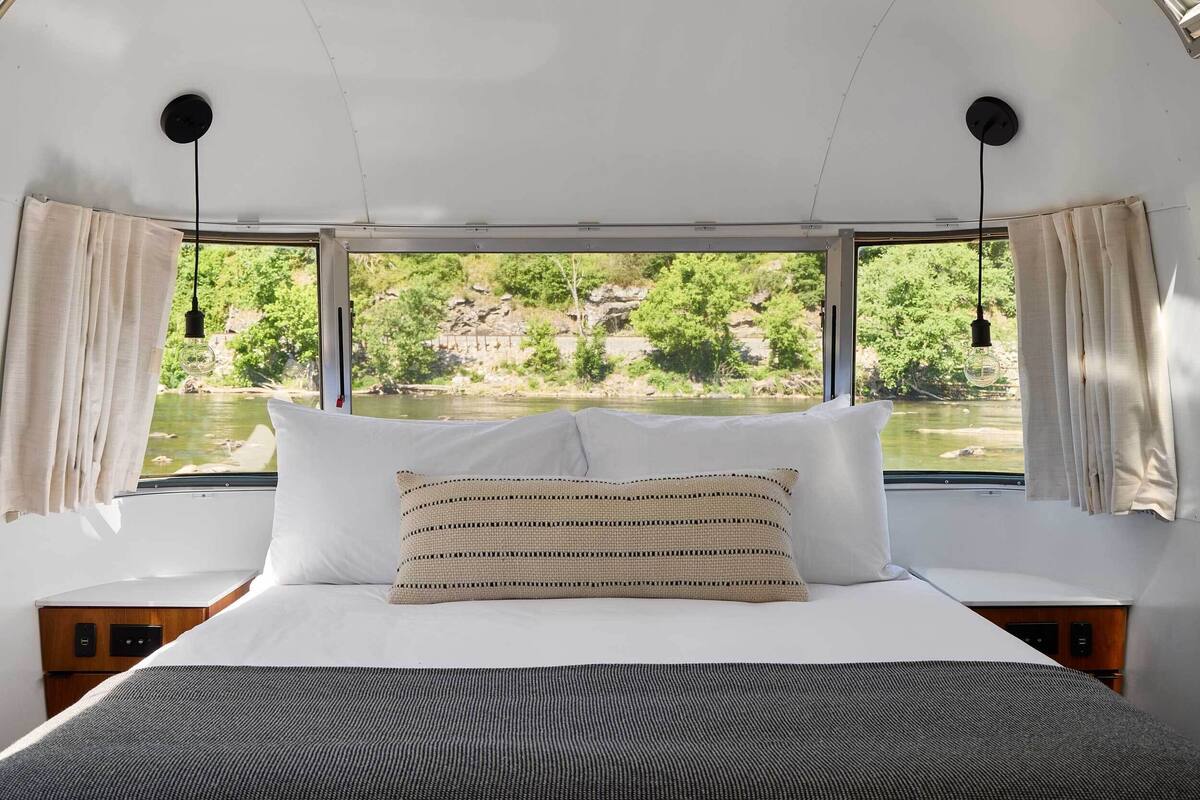 Una cama con almohadas blancas y a rayas, vista a un río y vegetación, y una ventana con cortinas.