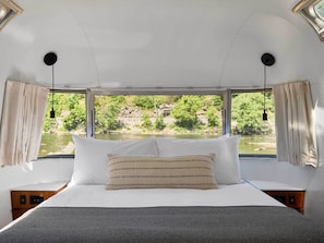 A bed with white and striped pillows, a view of a river and greenery, and a window with curtains.