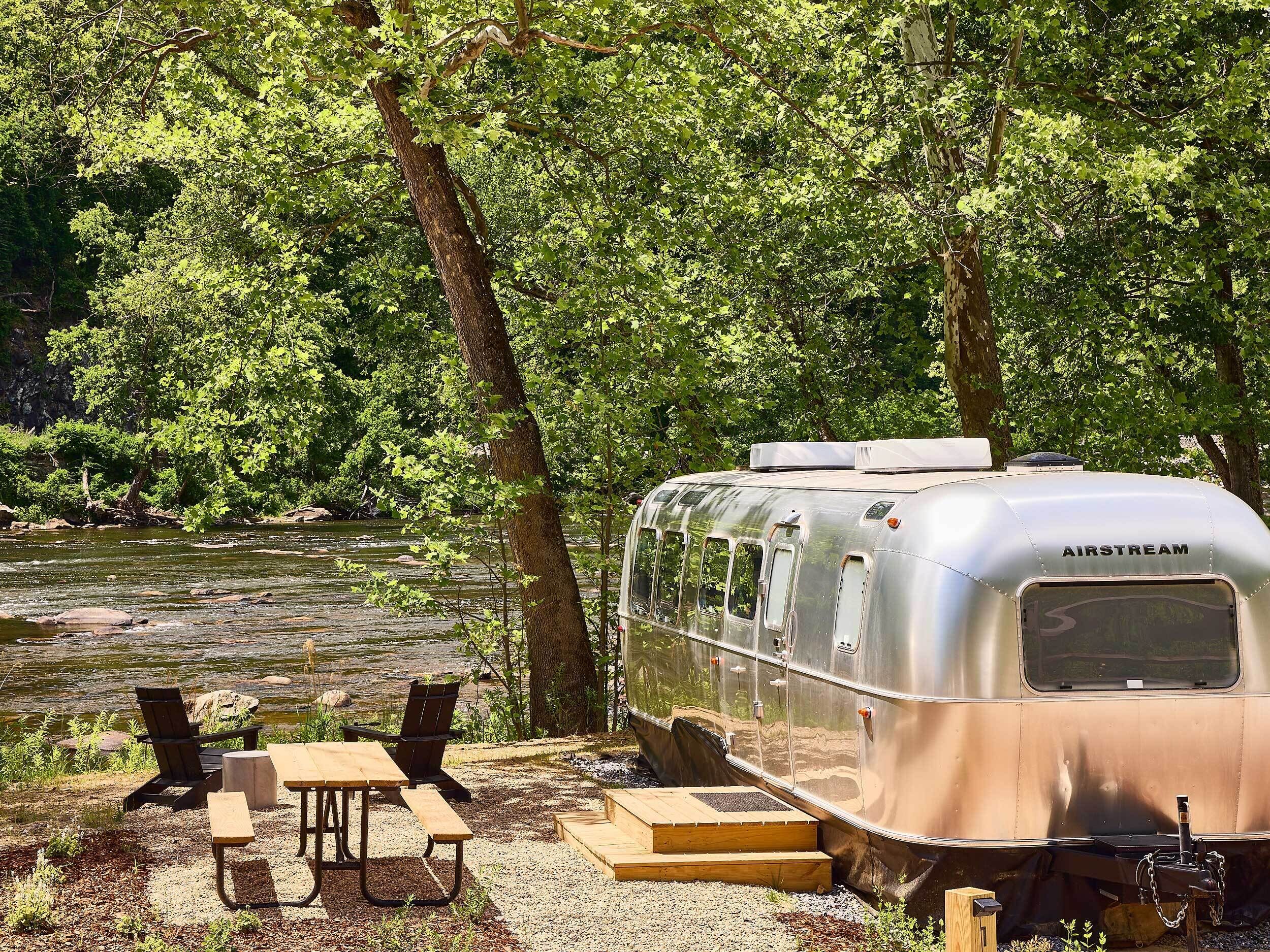 A silver AIRSTREAM trailer with a picnic table and chairs, set up by a river.