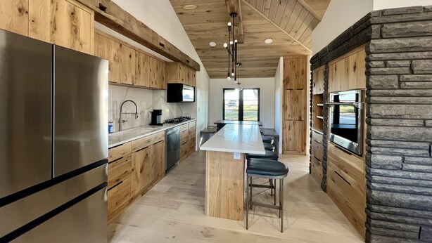 Une cuisine moderne avec des meubles en bois, un réfrigérateur en acier inoxydable, un îlot central doté de tabourets de bar et un mur d’accent en pierre.