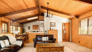 A cozy cabin interior with a wooden ceiling, a living area with a sofa and coffee table, a kitchen area, and a wooden door.
