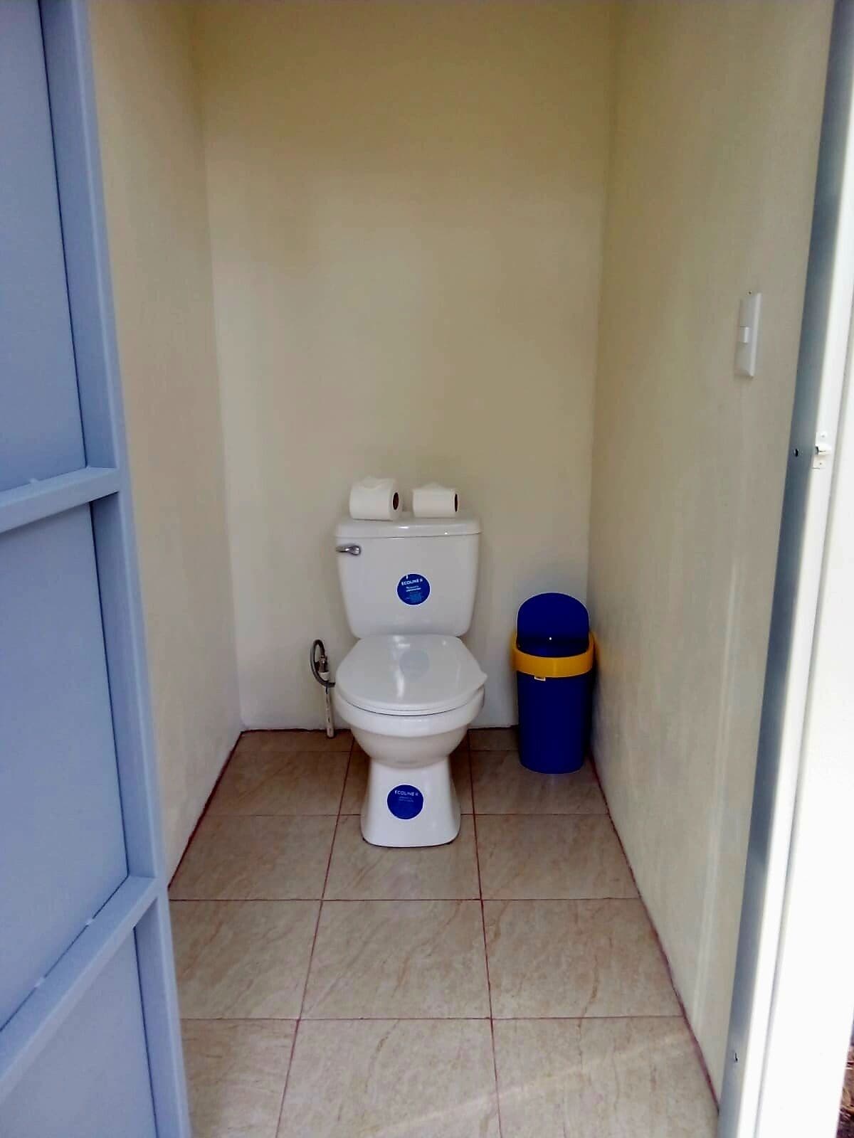 Un baño pequeño y estrecho con un inodoro blanco, un cubo de basura azul y suelo de baldosas.