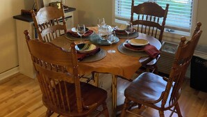 A dining area with a wooden table set for four, surrounded by wooden chairs. A chandelier hangs above, and a framed picture is on the wall.