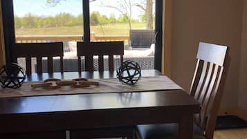 A dining area with a wooden table, chairs, and a bench. There is a large window with a view of trees and a grassy field.
